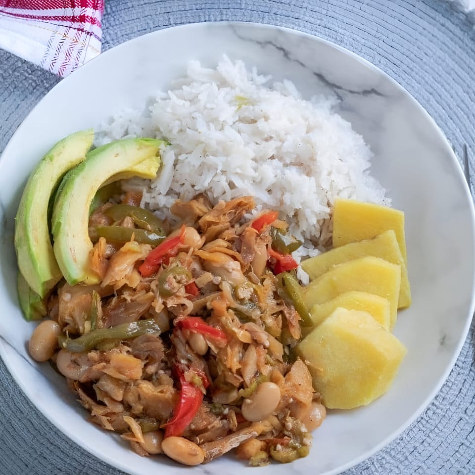 A Jamaican dish with rice, ackee and saltfish, sliced avocado, and boiled yellow yam on a white plate.